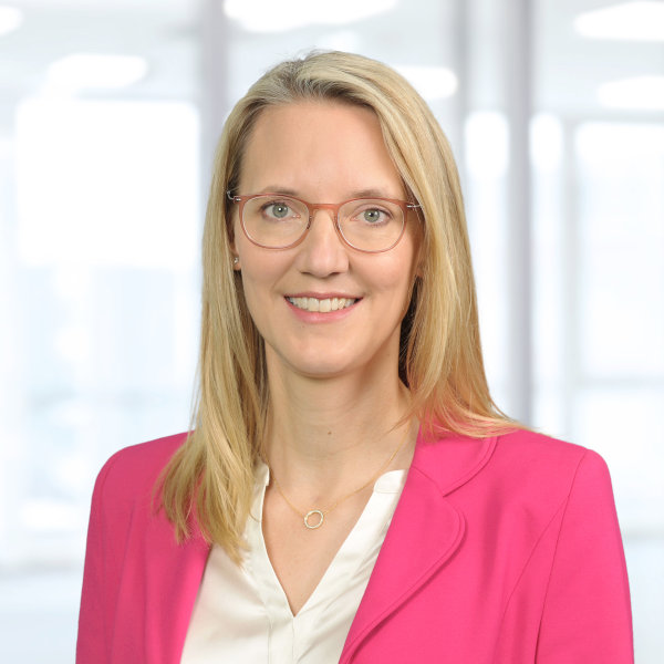 A woman wearing a pink blazer and a white blouse stands in a well-lit office environment.
