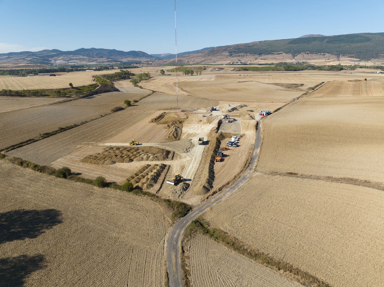 An aerial view of a construction site surrounded by fields, featuring heavy machinery and vehicles on a dirt road.