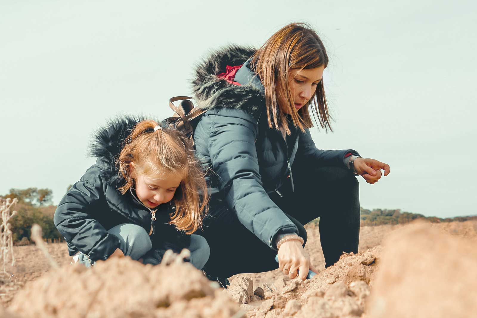 Una mujer y una niña se arrodillan en un campo, examinando la tierra mientras llevan abrigos en un día despejado.