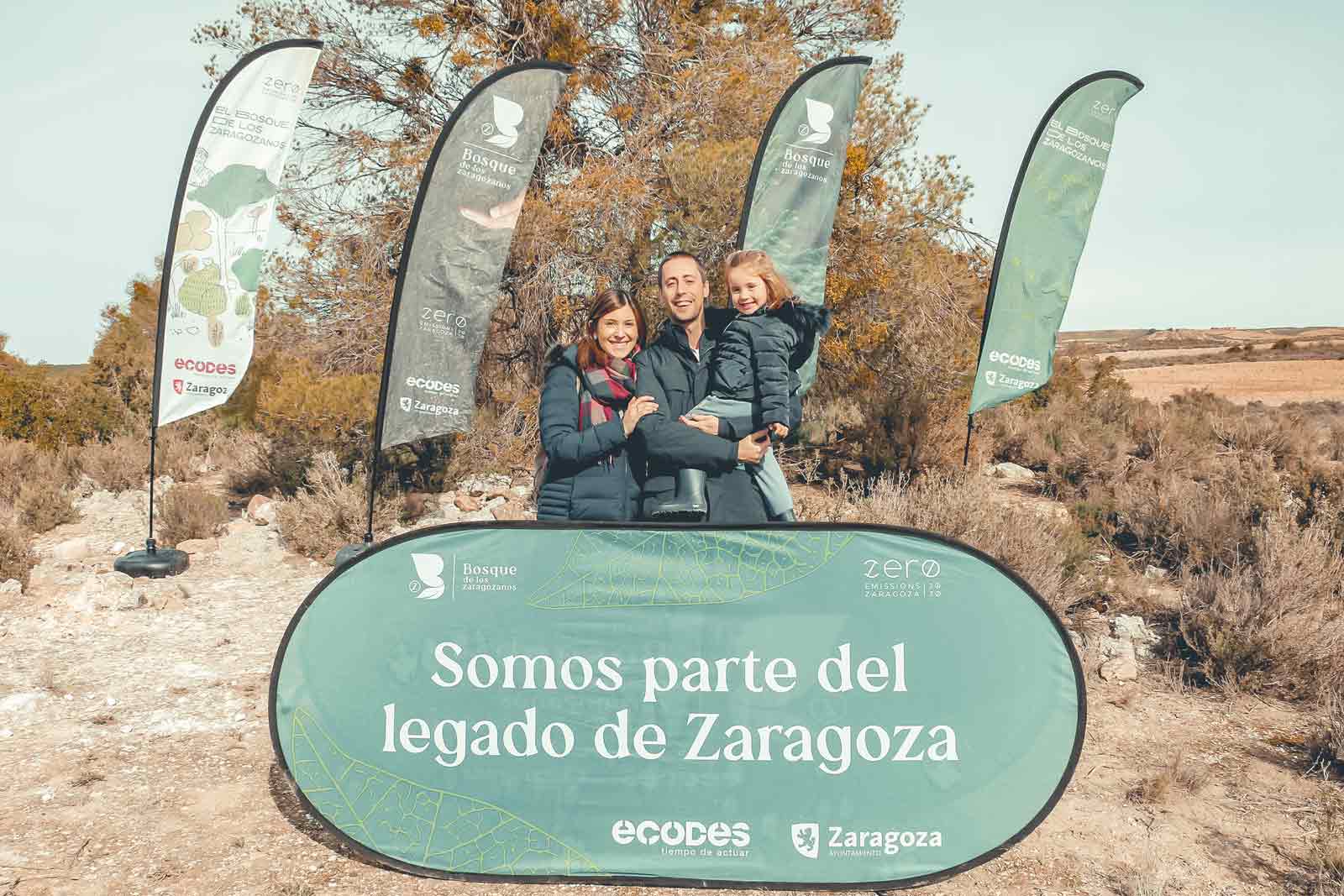 Una familia posa frente a un gran cartel en Zaragoza, promoviendo iniciativas ecológicas con banderas al fondo.