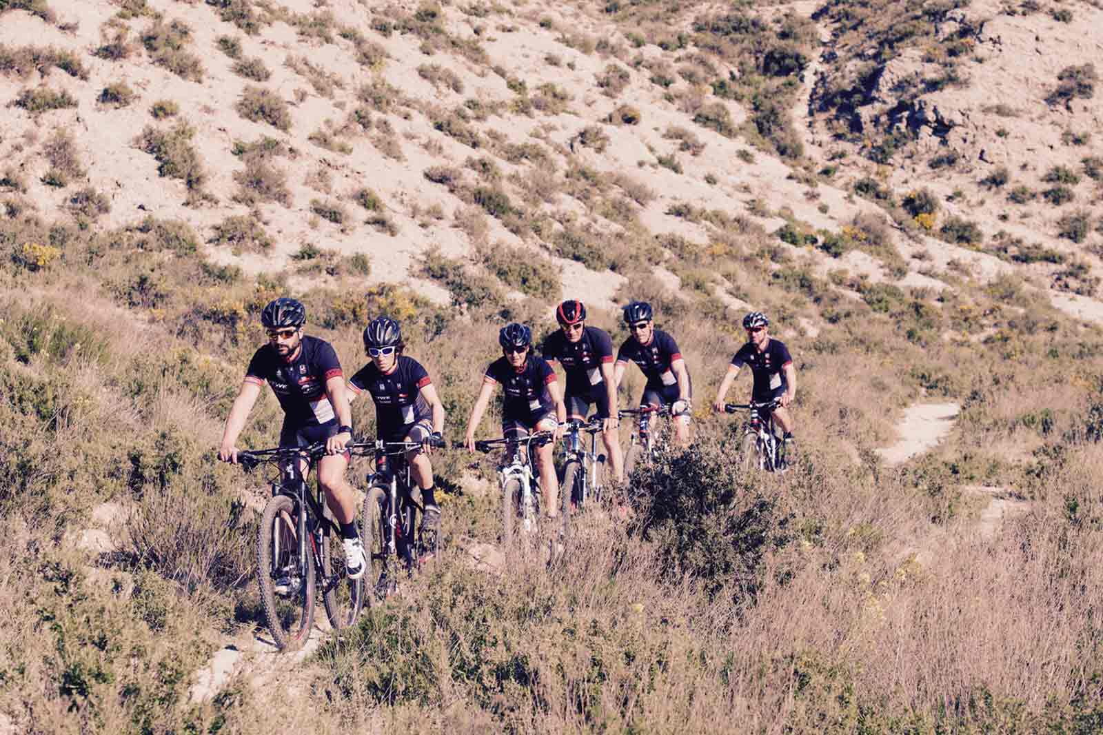 Un grupo de ciclistas montando en un camino de tierra a través de un paisaje seco y herboso con colinas al fondo.