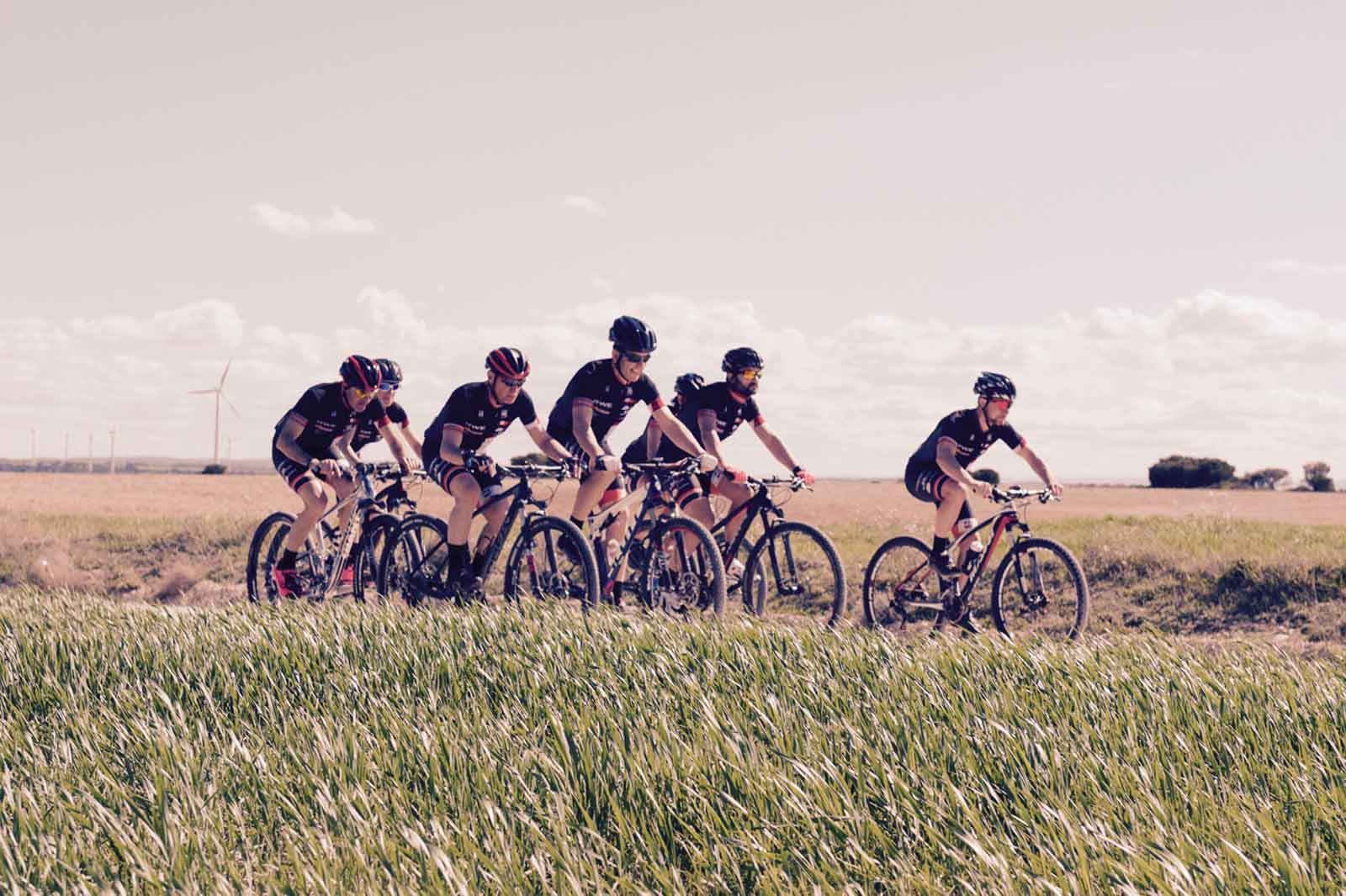 Una fila de ciclistas de montaña recorre un campo con aerogeneradores al fondo bajo un cielo parcialmente nublado.
