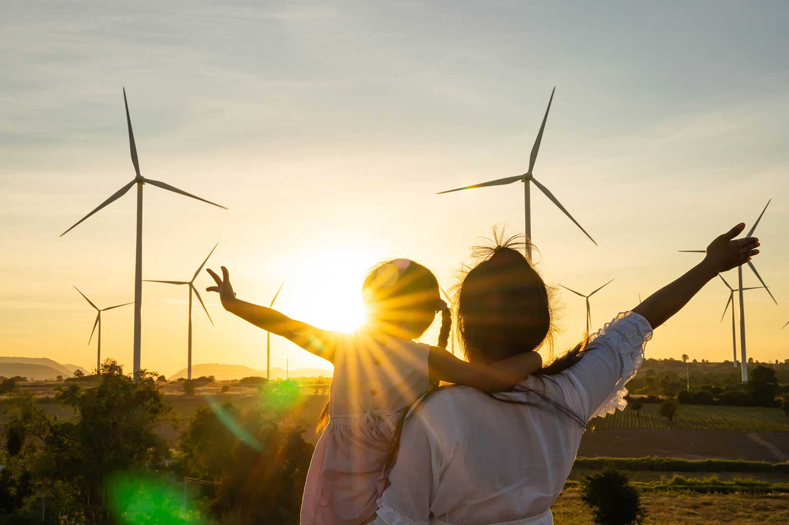 A mother and child are silhouetted against a sunset, surrounded by wind turbines in a scenic landscape.