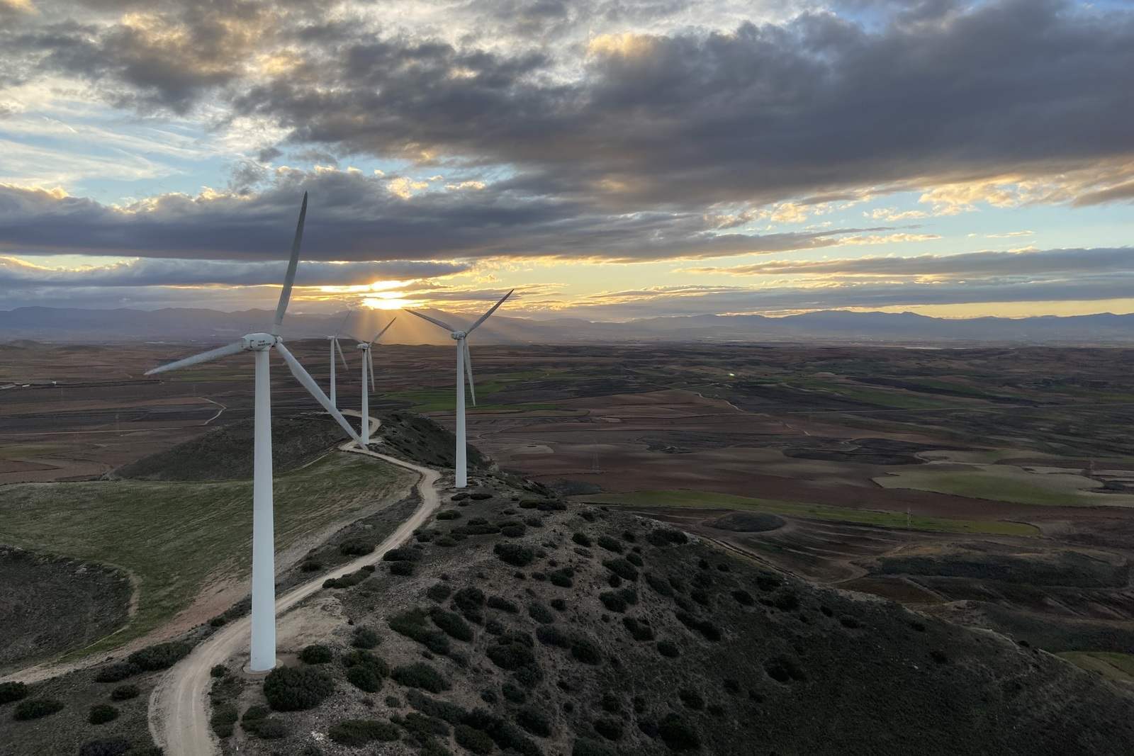 A scenic view of wind turbines on a hilltop during sunset, with dramatic clouds and expansive fields in the background.
