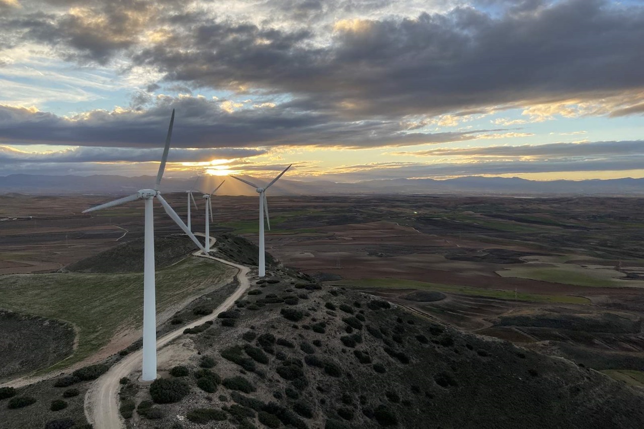 Una vista escénica de turbinas eólicas en una colina durante la puesta de sol, con nubes dramáticas y campos expansivos al fondo.