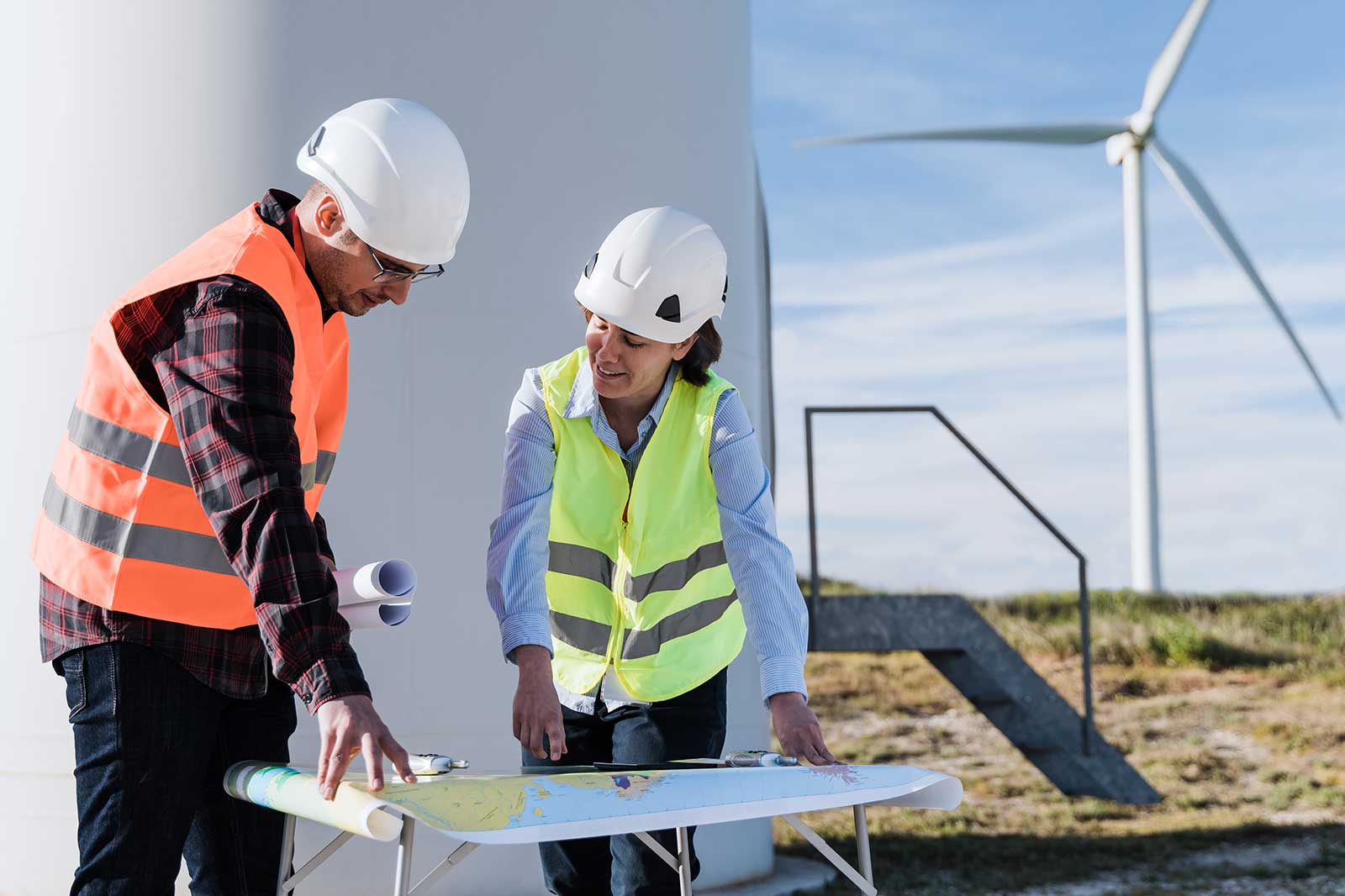 Two workers in safety gear discussing plans at a wind farm, with turbines visible in the background.