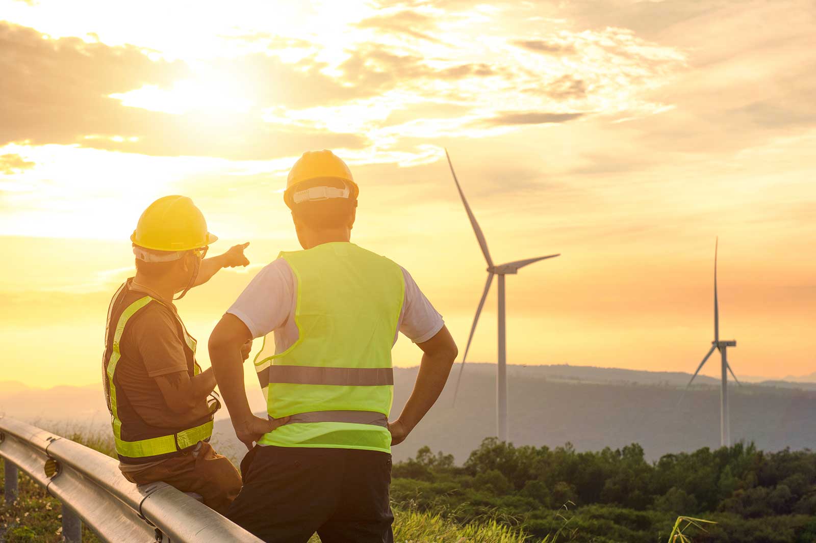 Two construction workers in safety gear observe wind turbines under a golden sunset, discussing their work.