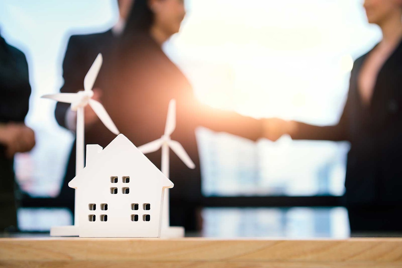 A model house with wind turbines is displayed as two professionals shake hands in a bright, modern office.