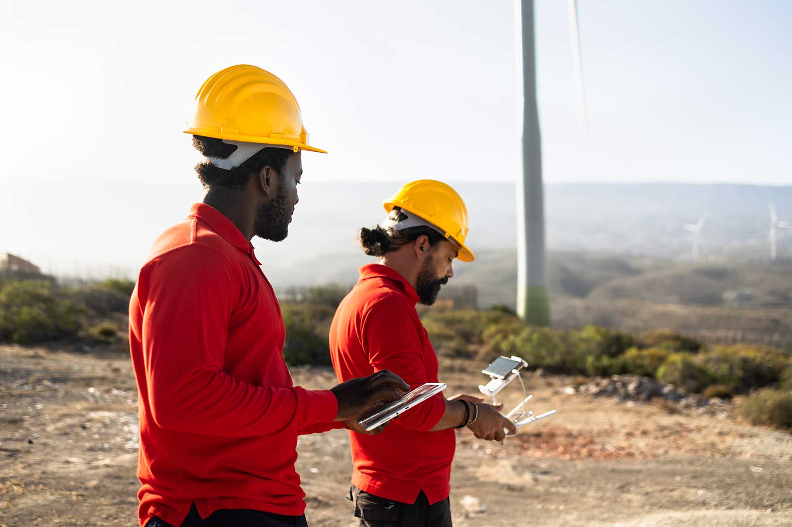 Two workers in yellow hard hats and red shirts stand outdoors, preparing to operate a drone near wind turbines.
