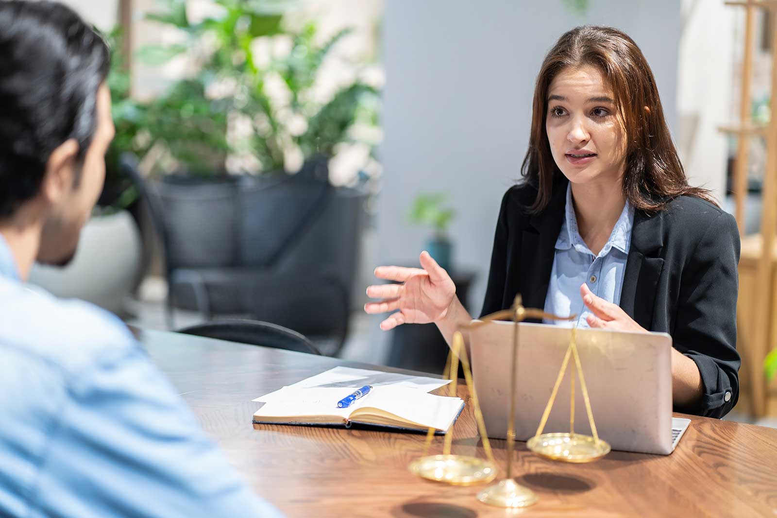 A woman in a black blazer discusses with a client at a wooden table, a laptop and scales of justice are visible.