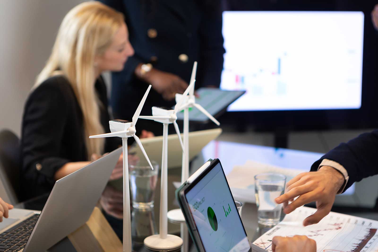 A meeting room with wind turbine models, laptops, and charts displayed on a screen.