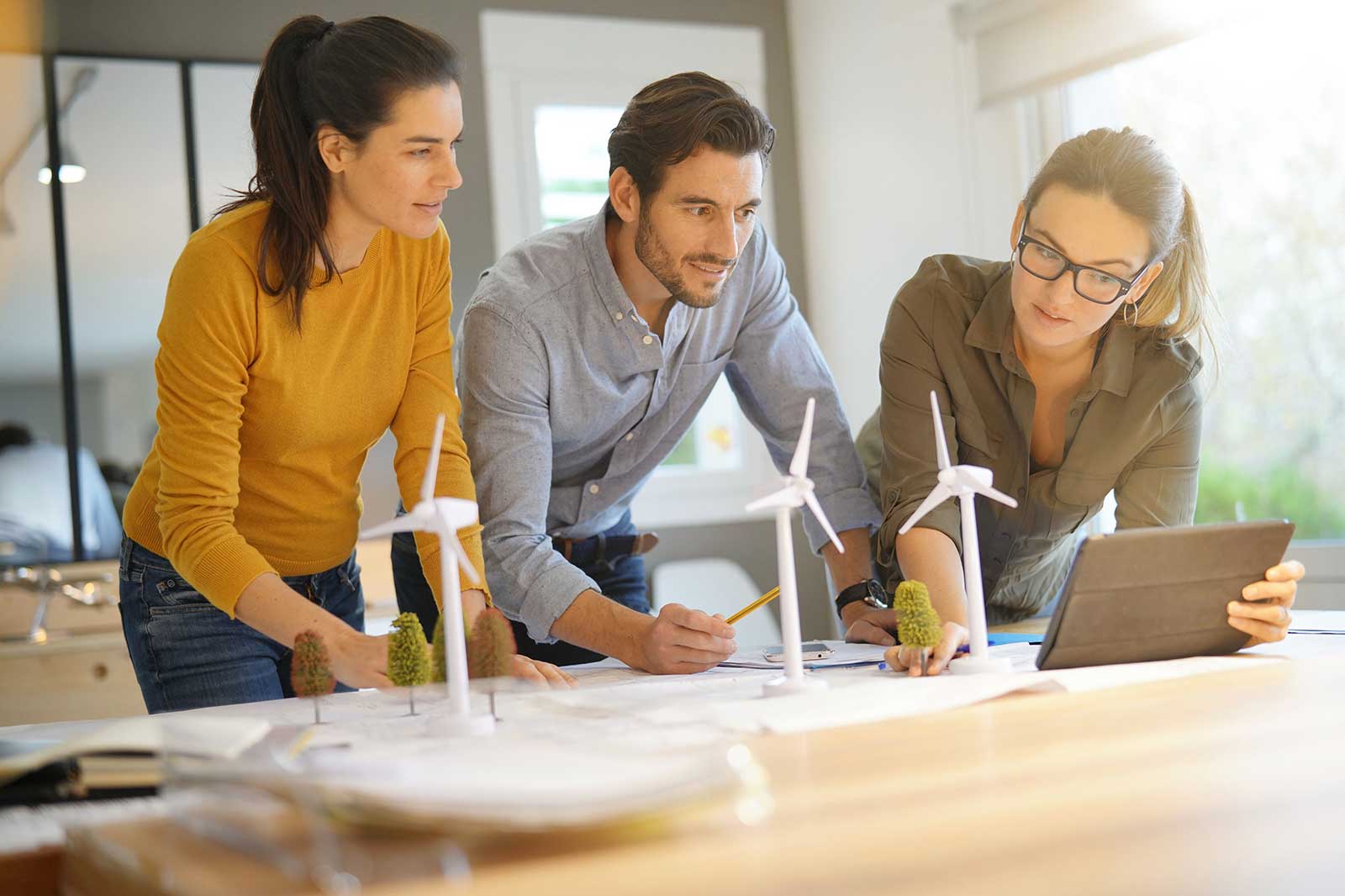 Three individuals collaborate on a project with wind turbine models and a tablet on a table.
