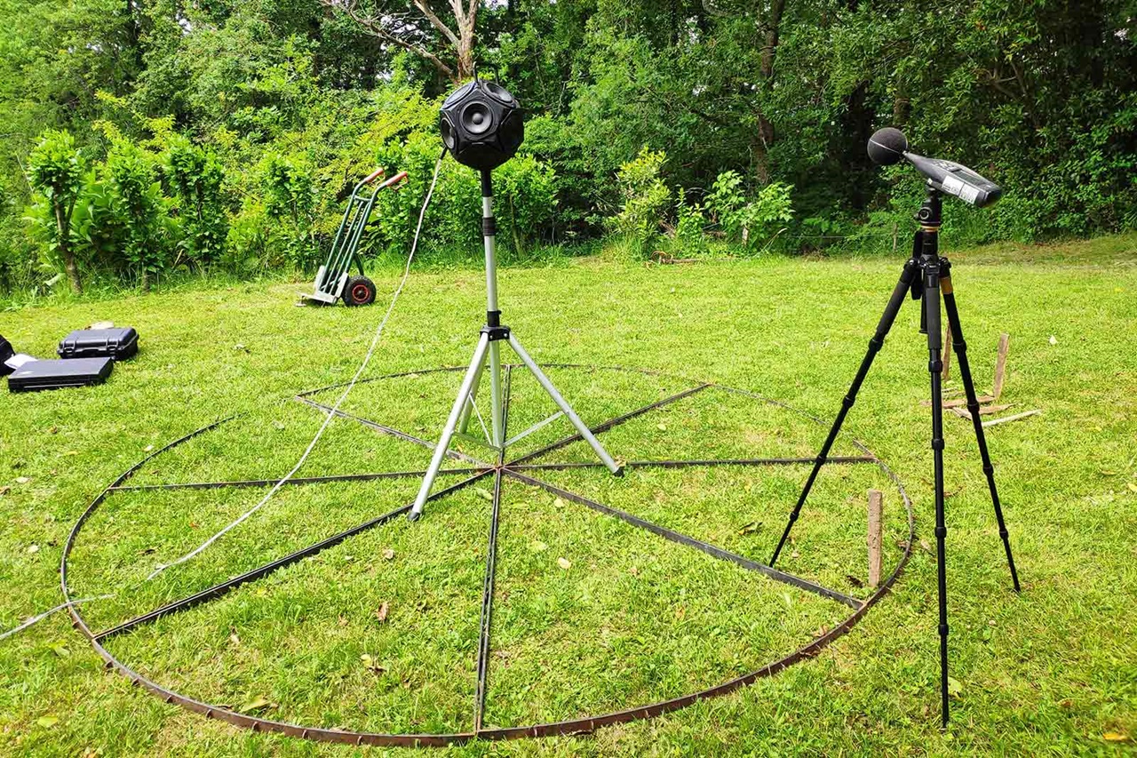 A microphone on a tripod next to a spherical audio device in a grassy area, surrounded by metal circle and equipment cases.