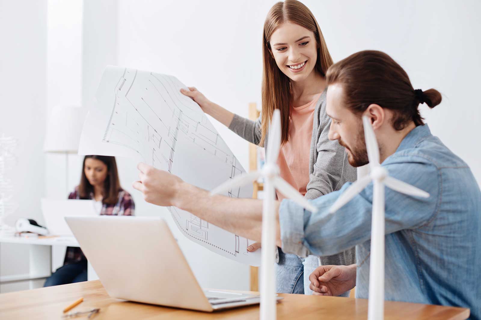 Two individuals discuss plans with wind turbines in foreground; a third person works at a laptop in the background.