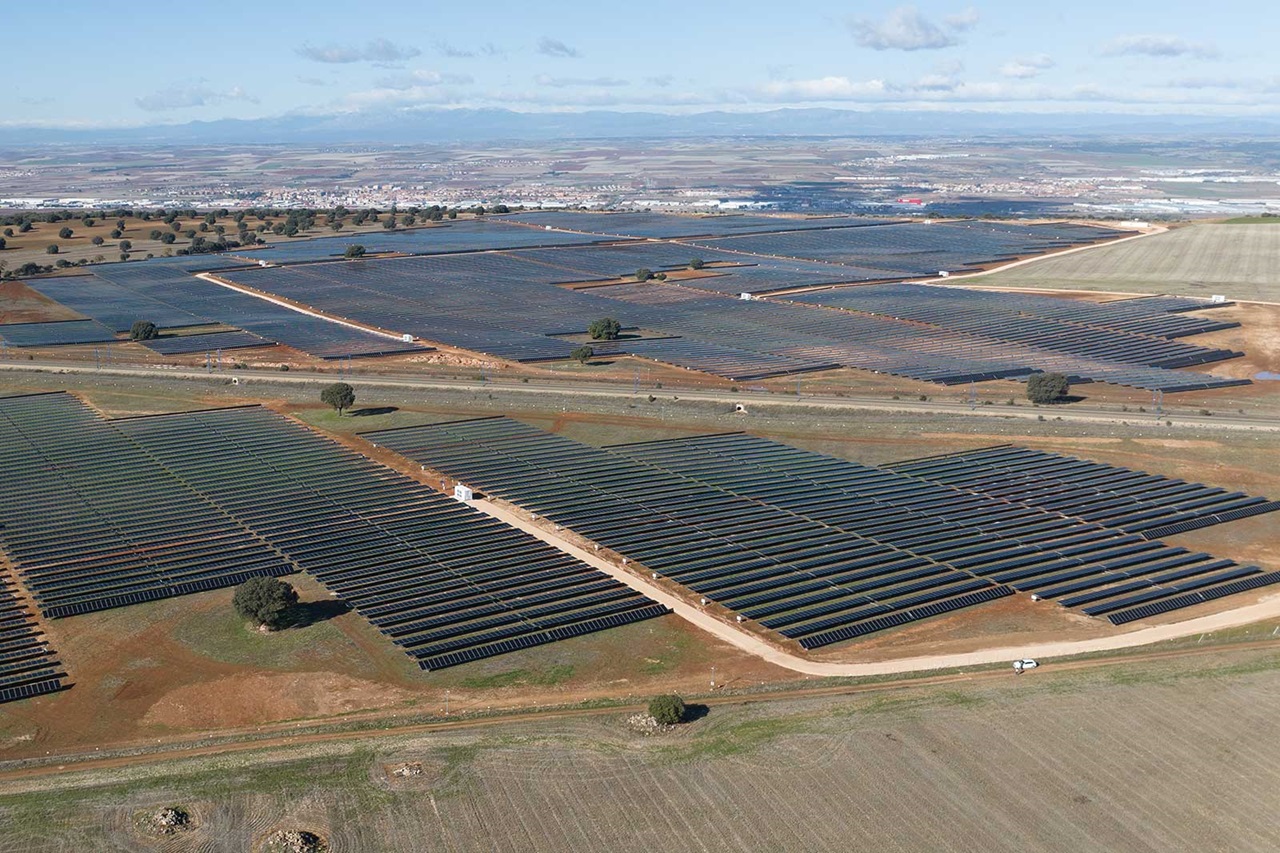 Aerial view of a large solar farm with rows of solar panels, surrounded by fields and distant mountains under a clear blue sky.