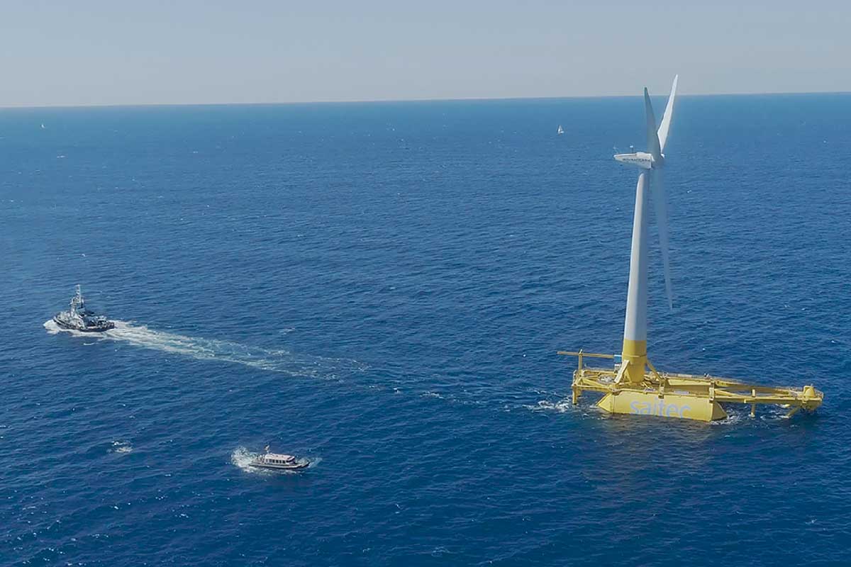 A view of a floating wind turbine in the ocean, with a tugboat and a smaller vessel nearby.