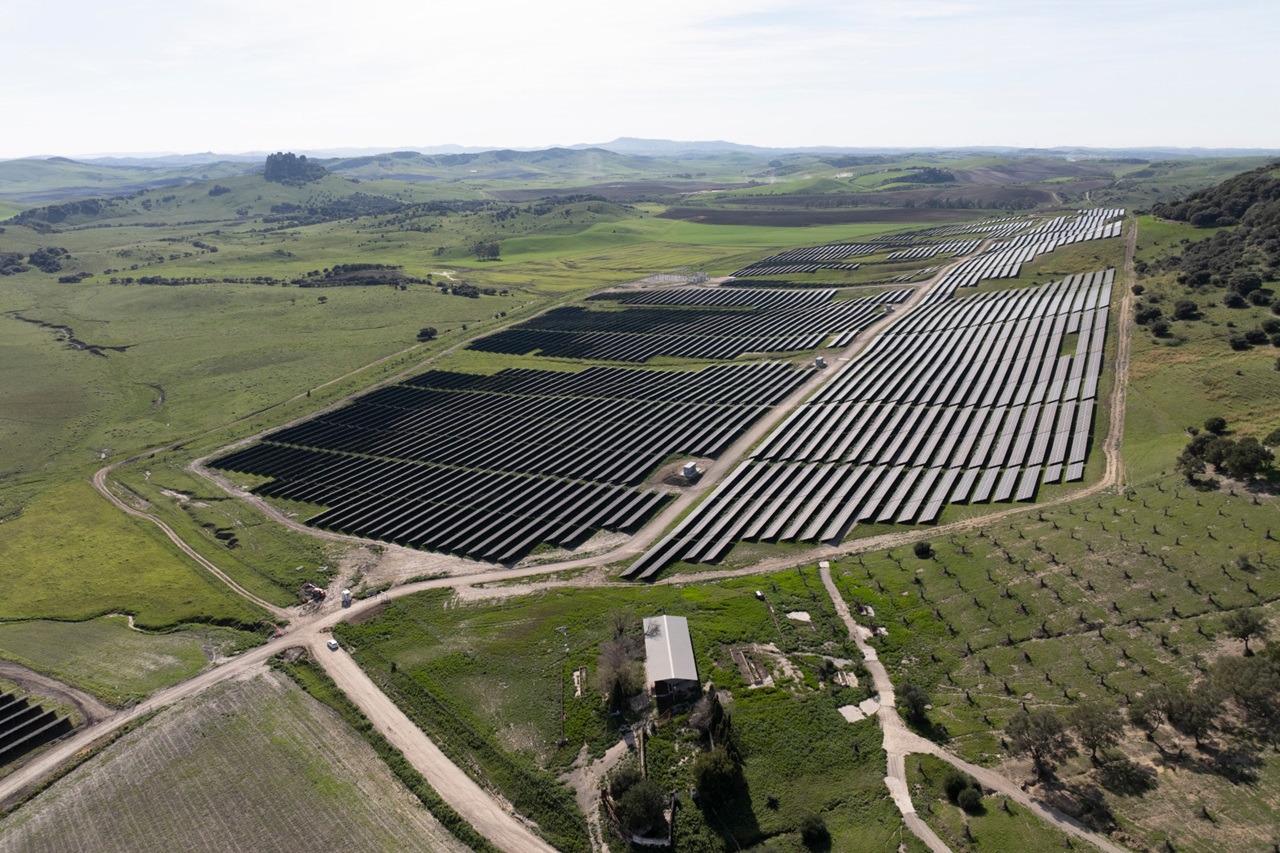 Una gran granja solar con filas de paneles solares en tierras verdes, rodeada de colinas y árboles.