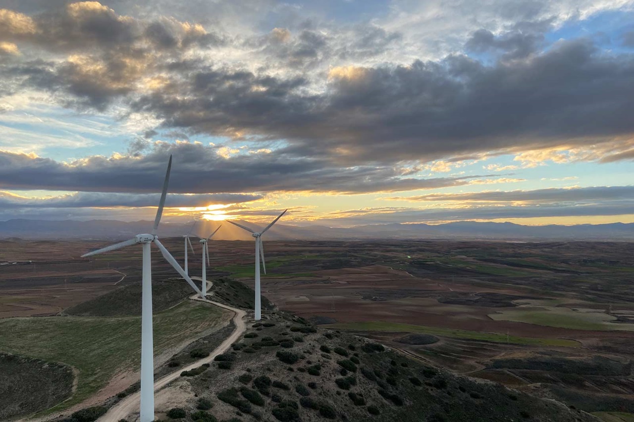 Tres aerogeneradores blancos en la cresta de una colina dominan el primer plano, con campos marrones y verdes y un cielo dramático al atardecer.