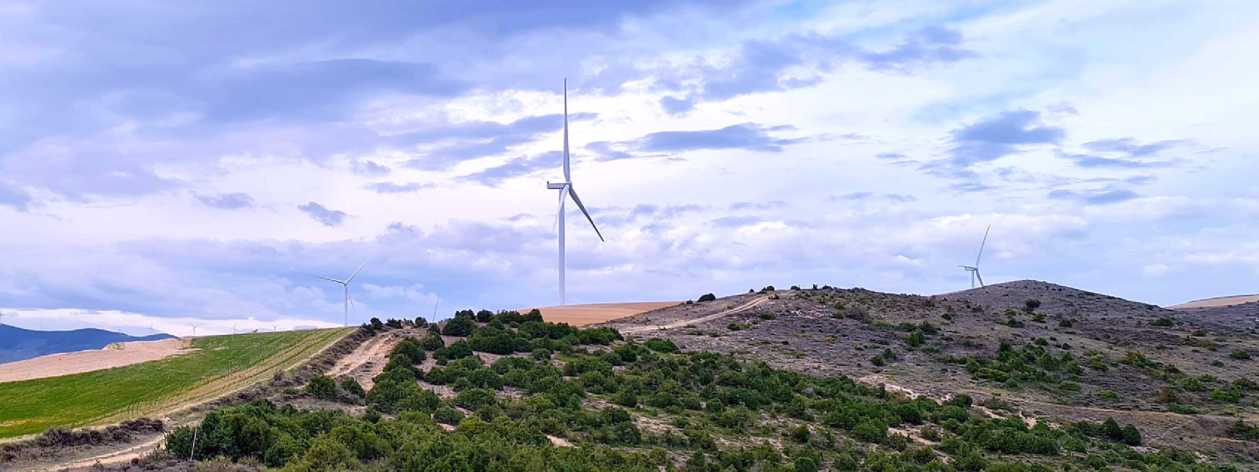 A landscape featuring hills, wind turbines, and a cloudy sky. The foreground showcases green fields and shrubs.
