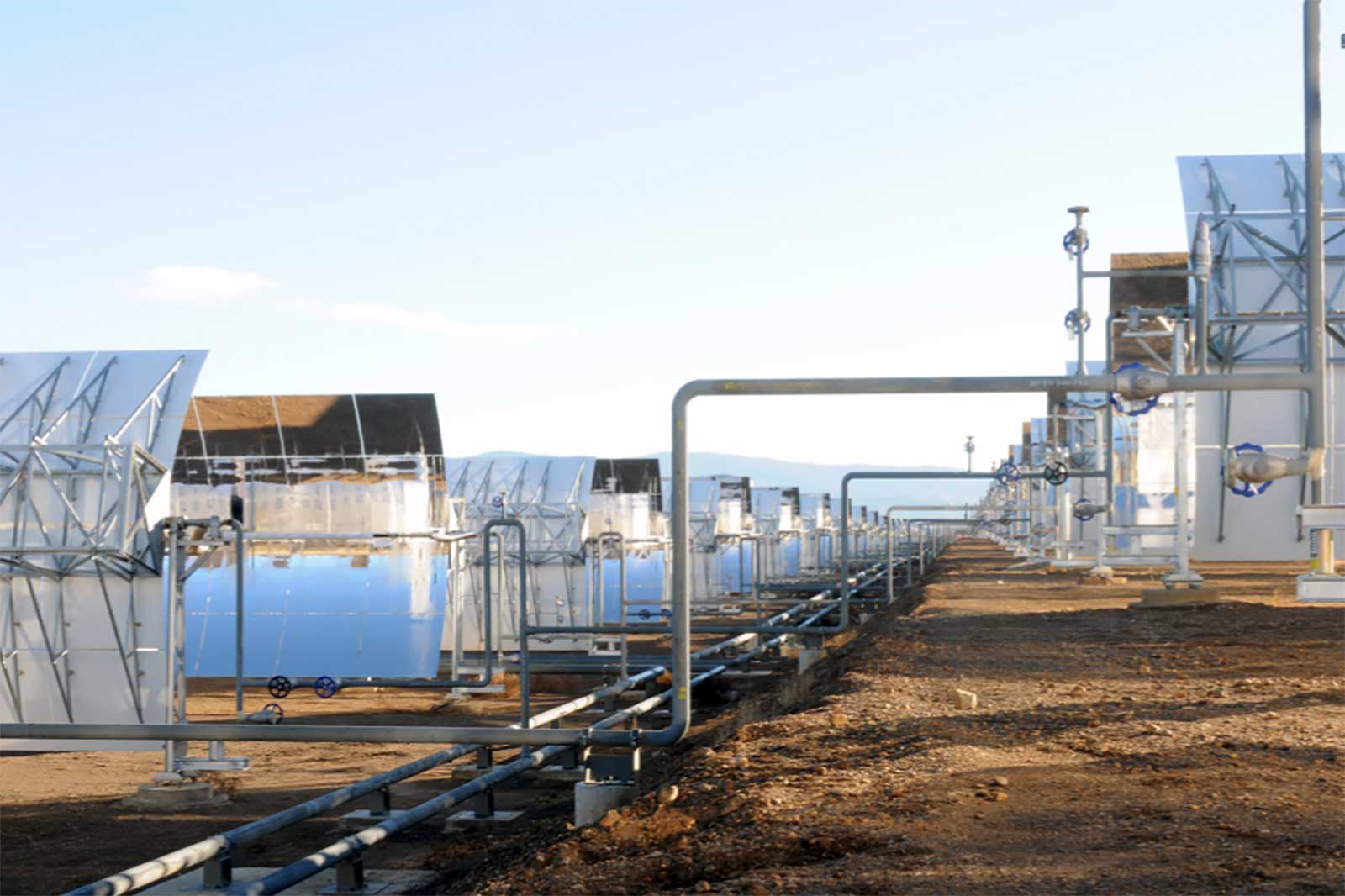 A row of solar panels reflecting sunlight with metal pipes and valves in a renewable energy facility.