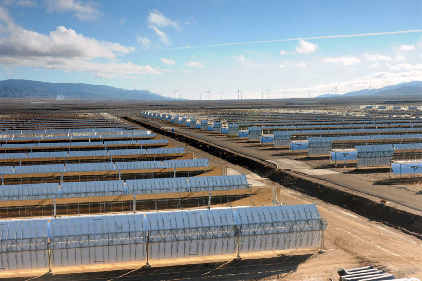A vast solar farm with numerous reflective panels stretching across the landscape, under a clear blue sky and distant wind turbines.