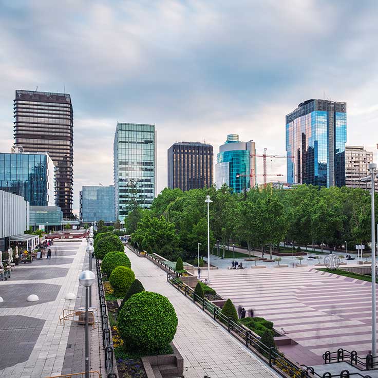 A modern urban landscape featuring glass buildings and landscaped walkways, with greenery and a cloudy sky.
