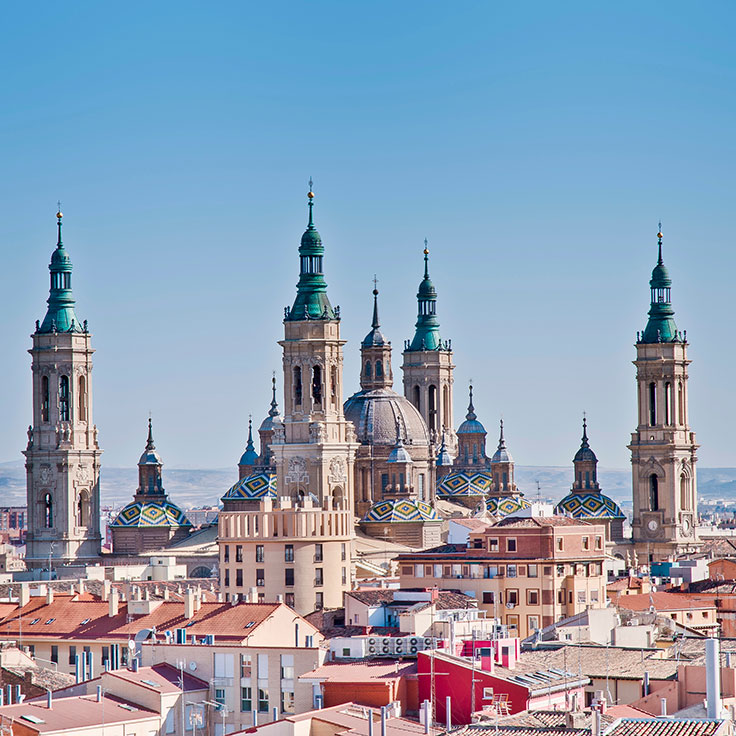 A panoramic view of tall church towers and domes against a clear blue sky in a cityscape with terracotta rooftops.