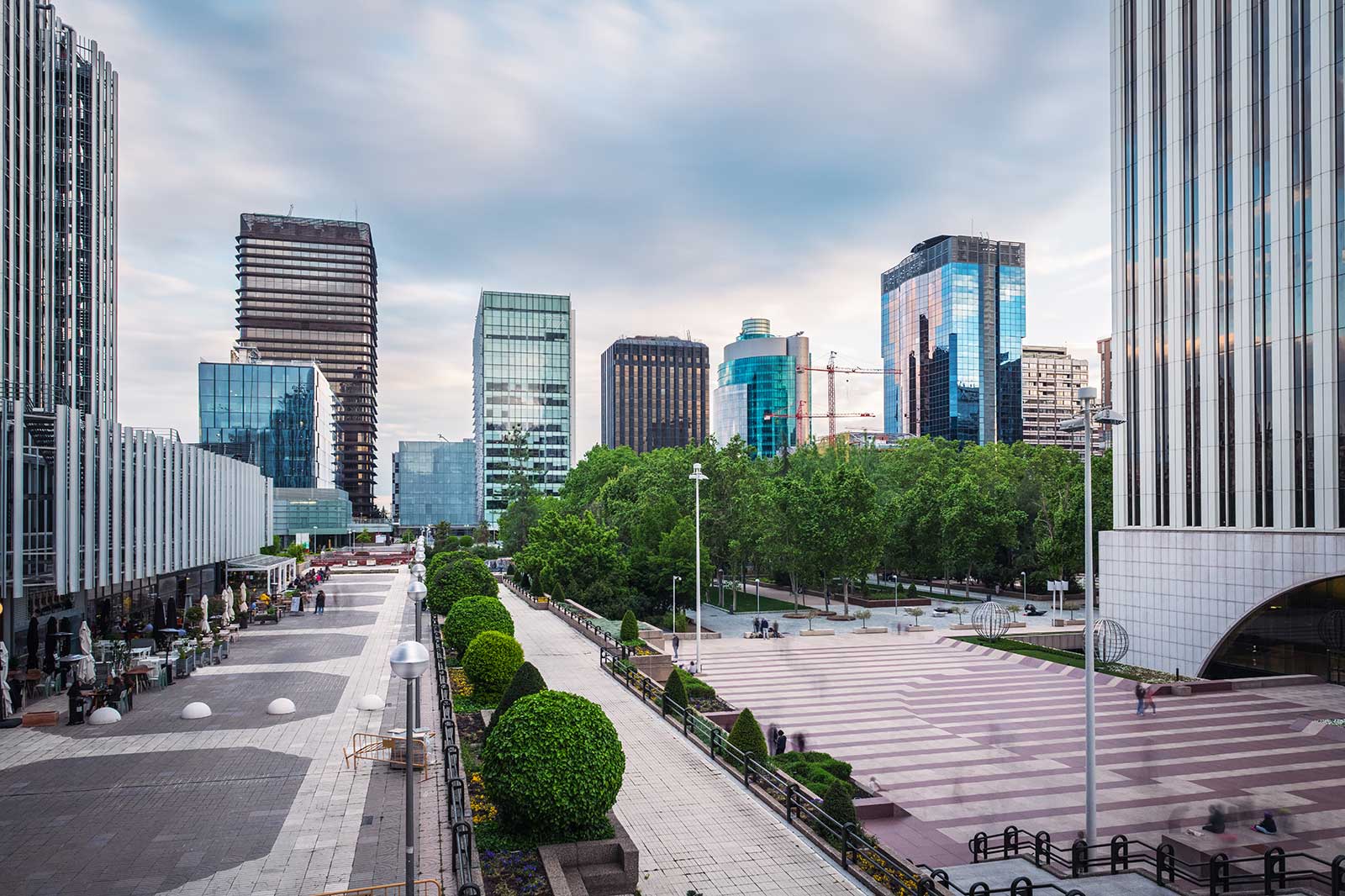 Un paisaje urbano moderno con rascacielos de cristal, vegetación y una plaza peatonal bajo un cielo nublado.