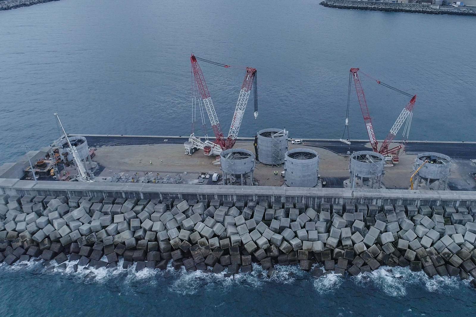 View of a harbour facility with cranes, cylinders, and a concrete wall by the water.