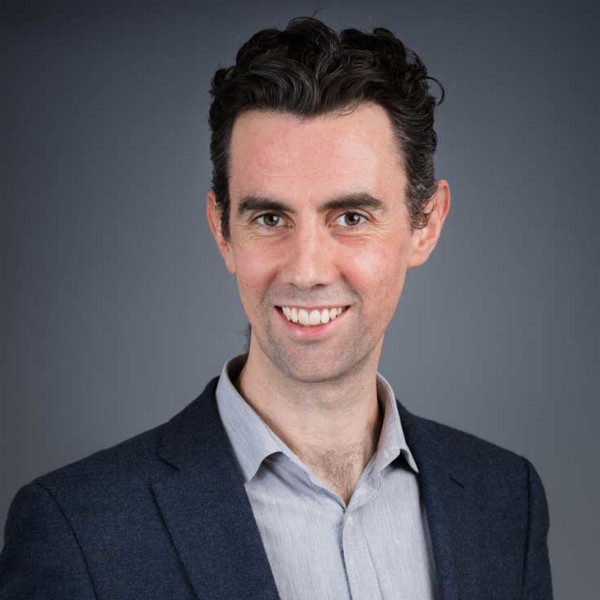 A professionally dressed man with curly hair, wearing a navy blazer and grey shirt, set against a plain background, portrait of Chris Willow.