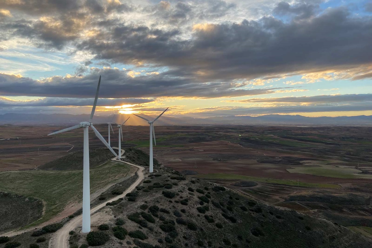 Wind turbines stand on a hill against a dramatic sky during sunset, overlooking a vast landscape of fields.