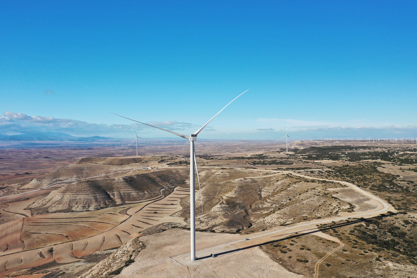 Una turbina eólica se encuentra en un terreno montañoso con suaves ondulaciones de tierra y un cielo azul claro de fondo.