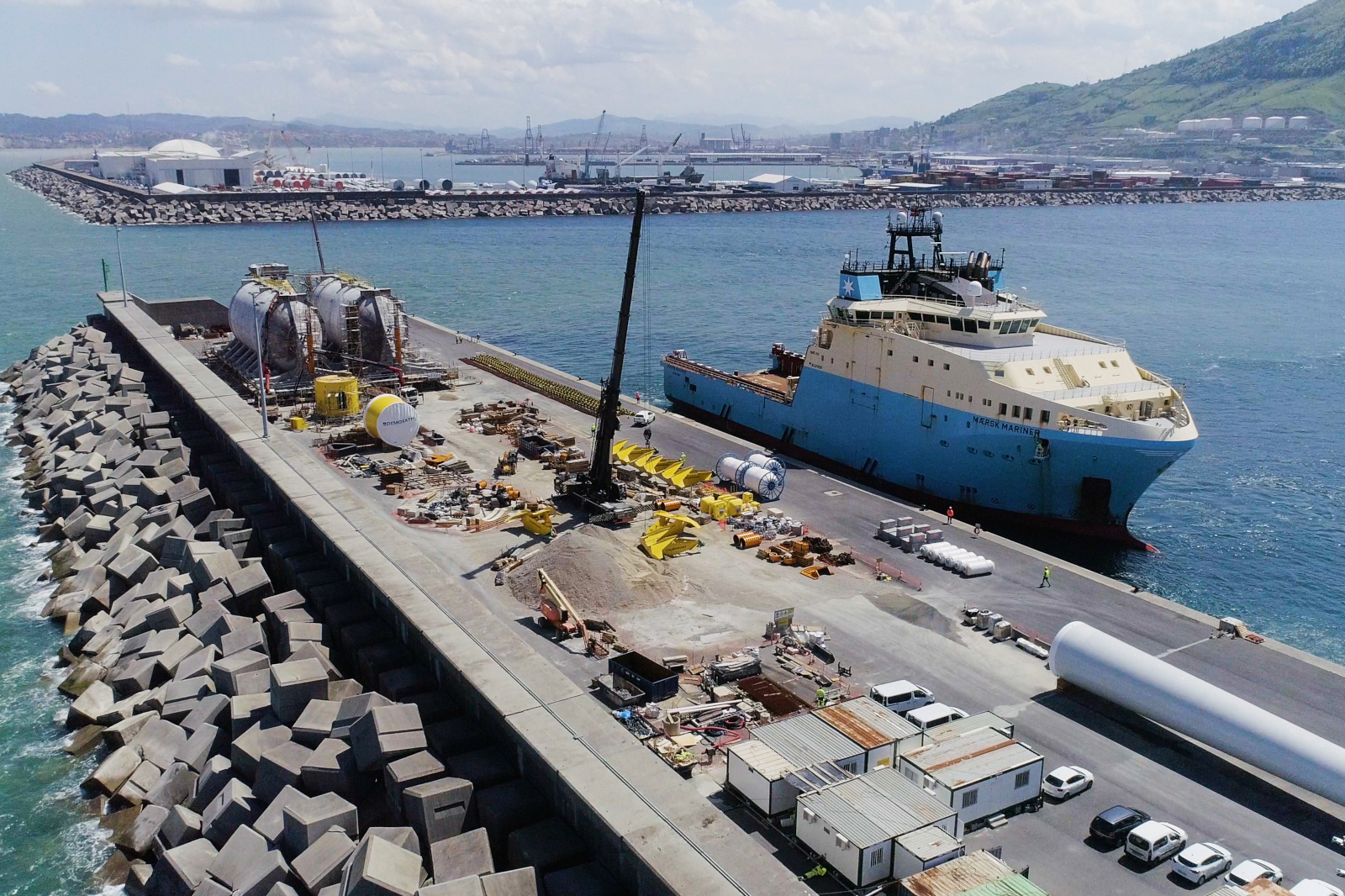 A port scene featuring a large blue vessel docked at a construction site with cranes, machinery, and various materials.