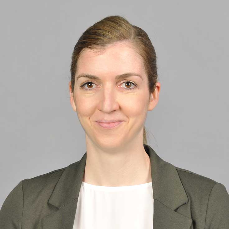 A professional woman wearing a dark blazer and white blouse against a neutral grey background. portrait of Sinje Vogelsang.